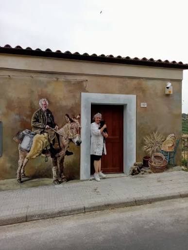 Murale di Pina Monne con una donna sulla porta e un uomo a cavallo di un asino
